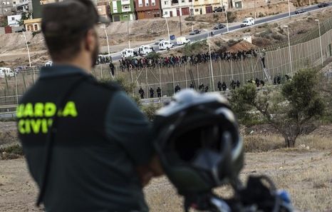 Un guardia civil vigila la frontera de Melilla, sobre cuya valla permanecen numerosos inmigrantes.
